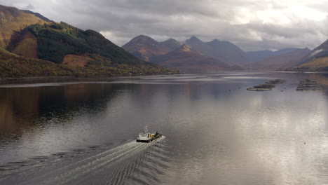 An-aerial-view-of-a-fishing-boat-heading-to-the-Loch-Duich-fish-farm-on-a-cloudy-day,-Northwest-Highlands-of-Scotland,-Glen-Shiel