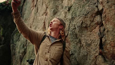Hiker-With-Long-Blond-Hair-Rising-His-Phone-To-Take-Selfie,-Rocky-Background,-Vogelberg