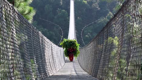 A-Nepali-person-carrying-a-heavy-load-and-walking-across-a-suspension-bridge-over-a-valley-in-Nepal