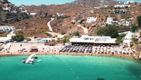 Super-Paradiesischer-Strand-In-Mykonos-Griechenland-Mit-Luftaufnahme-Eines-Sandstrandes-Mit-Menschen,-Resorts,-Restaurants-Und-Privaten-Villen-Auf-Den-Hügeln