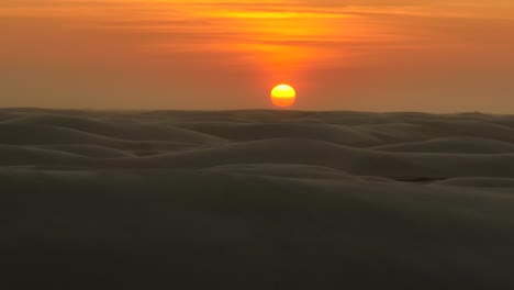 Low-sunset-aerial-of-sun-orb-in-deep-orange-sky-over-vast-undulating-sand-dunes