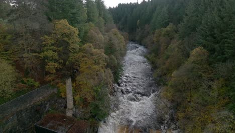 Drone-footage-reverses-down-a-fast-flowing-river-and-waterfall-surrounded-by-old-buildings-and-a-forest-of-broadleaf-and-coniferous-trees