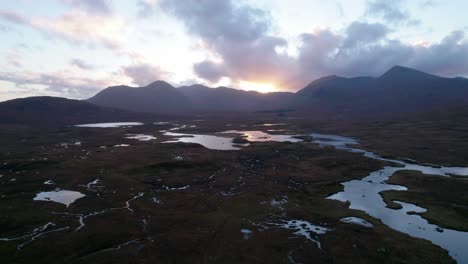 A-drone-slowly-reverses-high-above-a-peatland-landscape-of-peat-bogs-amongst-a-mosaic-of-fresh-water-lochs-looking-towards-mountains-on-the-horizon-at-sunset