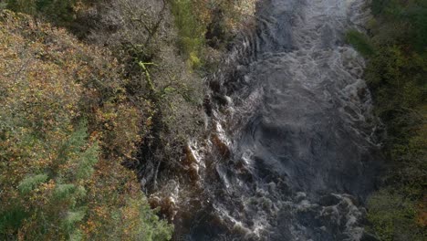 A-drone-flying-backwards-directly-over-a-fast-flowing-river-and-waterfall-tilts-to-reveal-a-forest-of-autumn-coloured-trees-and-blue-sky