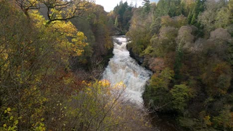 Drone-footage-of-a-cascading-waterfall-in-a-gorge-surrounded-by-colourful-trees-in-autumn,-slowly-twisting-to-to-reveal-the-waterfall-from-behind-the-trees