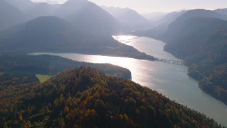 Autumnal-woodland-mountain-peak-aerial-view-towards-emerald-Sylvenstein-lake-reflecting-sunrise