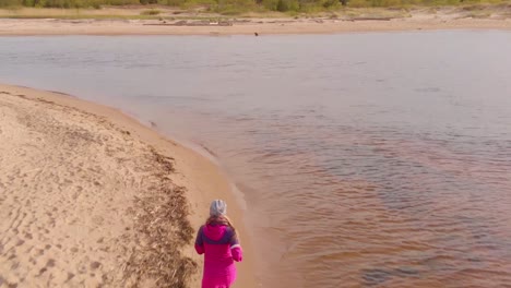 Young-girl-in-pink-jacket-jogging-on-the-seashore,-aerial-view
