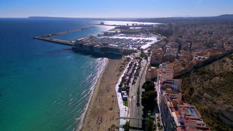 Aerial-drone-shot-of-playa-del-postiguet-in-Alicante-Spain-with-the-marina-and-small-sailing-ships-in-the-background