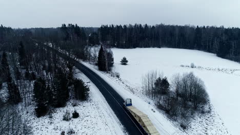 Aerial-View-Of-Winter-Snow-Landscape-With-Semi-Trailer-Transporting-Roof-Trusses-Along-Road