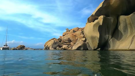 Half-underwater-panning-view-of-big-rocks-of-pink-granite-at-Cala-Della-Chiesa-bay-on-Lavezzi-island-with-luxury-boat-in-background