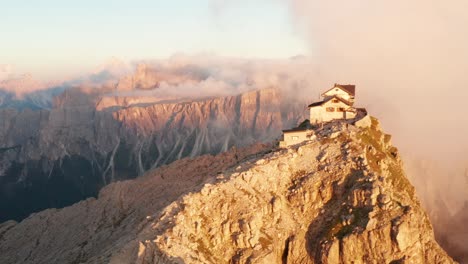 Striking-cliff-hut-perched-on-Nuvolau-mountain-peak-illuminated-by-sunset,-Italian-Dolomites,-aerial-trucking-shot