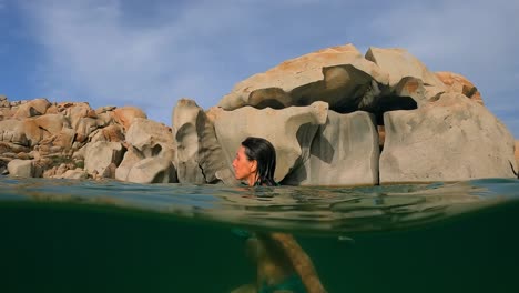 Half-underwater-scene-of-brunette-woman-in-bikini-swimming-in-calm-sea-water-of-Cala-Della-Chiesa-lagoon-with-eroded-granitic-rocks-in-background-on-Lavezzi-island-in-Corsica,-France