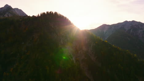 Golden-sunrays-emerging-behind-rocky-Tyrol-woodland-alps-mountain-range-silhouette-peaks-aerial-view-orbiting-right