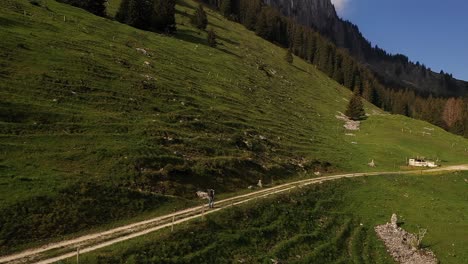 Aerial-drone-shot,-backpacker-hiking-on-mountain-cliffside-dirt-trail-during-summer