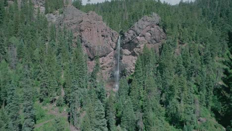 Distant-aerial-view-of-a-big-waterfall-in-a-rocky-mountain-with-a-lot-of-pine-trees