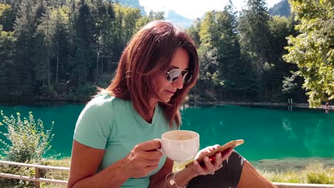 Sporty-and-attractive-older-woman-relaxing-by-a-lake-in-nature-after-exercising-in-summer,-on-the-phone,-drinking-coffee-and-having-a-quick-glance-at-the-camera