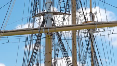 Tall-ship-rigging-gently-swaying,-backlit-against-a-blue-sky-with-puffy-clouds,-showing-three-masts-and-crow's-nest