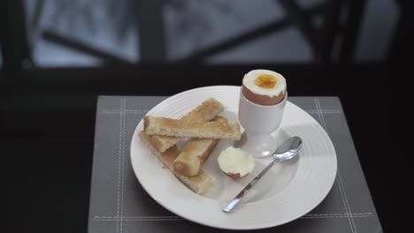 Breakfast-boiled-egg-in-an-eggcup-with-toast-on-plate,-black-table