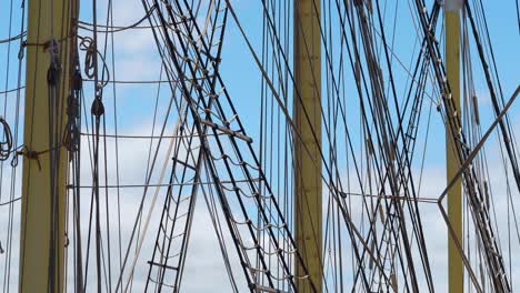 Tall-ship-rigging-gently-swaying,-backlit-against-a-blue-sky-with-puffy-clouds