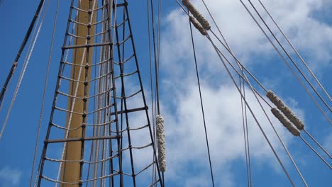 Tall-ship-rigging-gently-swaying,-frontlit-against-a-blue-sky-and-fast-moving-clouds