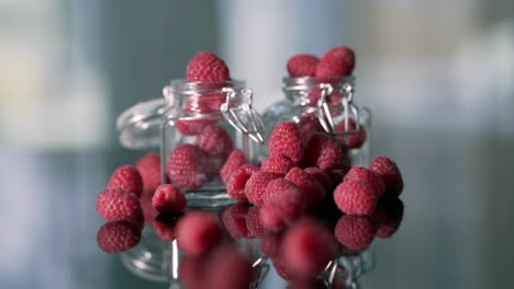 Beautiful-whole-raspberries-spilling-out-of-tiny-jam-pots-movement-from-black-table-to-raspberries