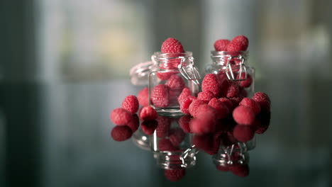 Red-raspberries-spilling-from-glass-pots-on-a-table,-garden-background-and-movement-from-left-to-right