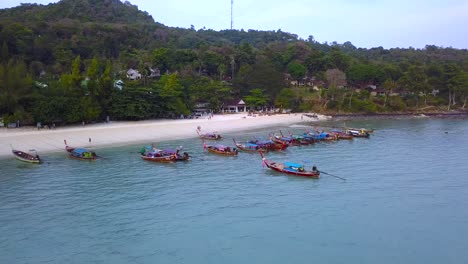 4K-AERIAL-FORWARD-VIEW-of-beach-with-long-boats-moored-in-Phi-Phi-Island-bay,-Phi-Phi-Don,-Thailand