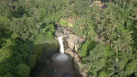 AERIAL-RISING-DOLLY-BACKWARD-VIEW-OF-A-CASCADE,-Tegenungan-Waterfall-in-Bali,-Indonesian,-with-surrounding-forest