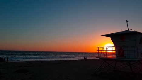 Slow-sideways-rising-up-sunset-shot-of-Lifeguard-house-:-tower-with-silhouettes-of-people-by-the-shore-at-San-Buenaventura-State-Beach-in-Ventura,-California,-United-States