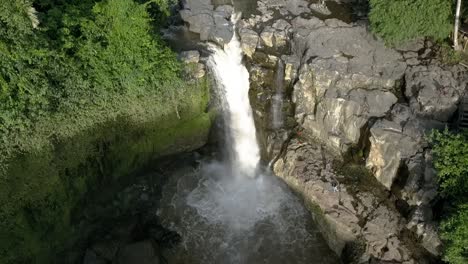 AERIAL-TILT-DOWN-OVER-WATERFALL-of-BALI-,-Tegenungan-Waterfall,-Indonesia,-with-surrounding-rocks