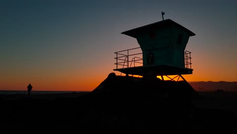 Slow-and-low-sideways-sunset-shot-of-Lifeguard-house-:-tower-with-silhouettes-of-people-by-the-shore-at-sunset-at-San-Buenaventura-State-Beach-in-Ventura,-California,-United-States