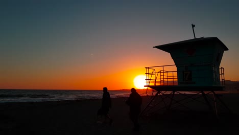 Slow-and-low-sideways-sunset-shot-of-Lifeguard-house-:-tower-with-silhouettes-of-people-passing-in-front-at-sunset-at-San-Buenaventura-State-Beach-in-Ventura,-California,-United-States