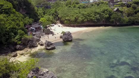 PADANG-PADANG-BEACH,-Low-flying-over-crystal-water-with-rocky-outcrop-on-sea