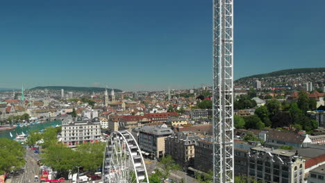 Aerial-drone-crane-shot-going-down-showing-amusement-park-Ferris-wheel-and-free-fall-tower-with-the-city-of-Zürich,-Switzerland-in-the-background-during-Zürichfest