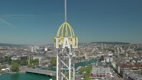 Aerial-drone-shot-flying-close-towards-amusement-park-free-fall-tower-with-the-city-of-Zürich,-Switzerland-in-the-background-during-Zürichfest
