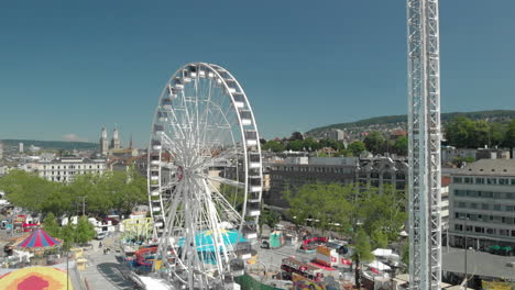 Aerial-drone-crane-shot-of-amusement-park-Ferris-wheel-and-free-fall-tower-with-the-city-and-lake-of-Zürich,-Switzerland-in-the-background-during-Zürichfest