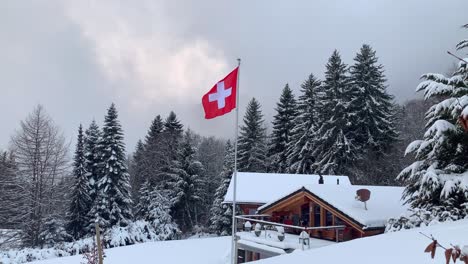 Bandera-Suiza-Sobrevolando-Los-Alpes-Suizos-En-Invierno-Con-Un-Chalet-Tradicional