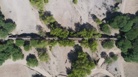 Group-of-friends-riding-bicycle-crossing-an-old-bridge-through-an-amazing-landscape,-cenital-drone-shot