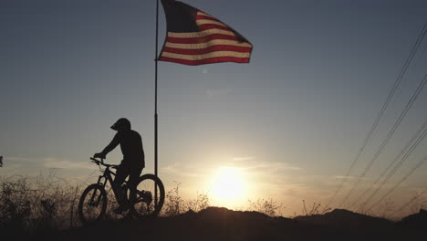 Ciclista-De-Montaña-Parado-Bajo-La-Bandera-Americana-Al-Atardecer-Con-Montañas-Al-Fondo