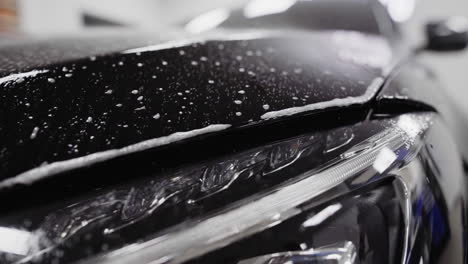 Slow-motion-pan-across-the-front-of-a-black-car-as-sudsy-spray-wets-the-hood-of-the-car