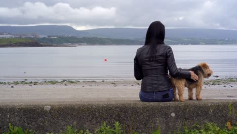 Hund-Und-Besitzerin-Sitzen-An-Der-Strandmauer-Und-Betrachten-Das-Leben-Mit-Blick-Auf-Das-Meer-Und-Die-Hügel