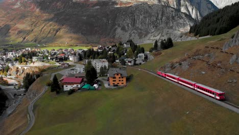 Aerial-View-of-Red-Swiss-Train-on-Railway-Arriving-to-Small-City-Under-Alps,-Switzerland