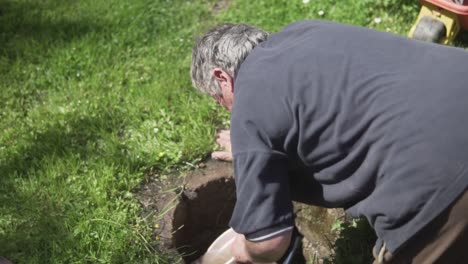 Slow-motion-back-shot-of-old-man-kneeling-and-pulling-out-water-from-a-ground-hole-using-a-bucket