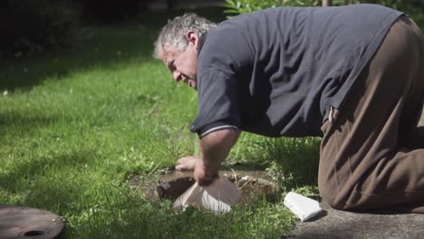 Slow-motion-shot-of-old-man-kneeling-and-pulling-out-water-from-a-ground-hole-using-a-bucket