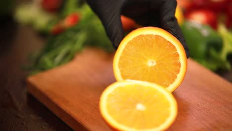 Juice-fresh-orange-cutting-by-large-knife-on-chopping-board-by-hands-wearing-black-gloves-with-vegetables-on-background