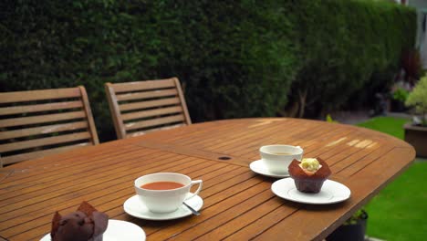 Tee-In-Porzellantassen-Auf-Holztisch-Frau-Bringt-Zwei-Muffins-Zum-Essen-Im-Sommergarten