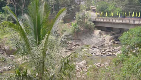View-of-the-river-moving-beneath-a-bridge-over-Nallathanni-river-in-munnar-hills-in-the-drier-season-of-the-year-with-dense-forest-around-and-yellow-flags-for-a-religious-celebration