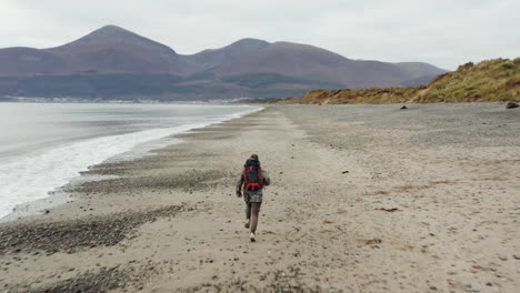 Backpaker-male-running-along-beach-escaping-towards-town-and-mountains-past-dunes