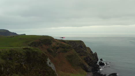 Drone-with-red-lights-flies-from-green-rocky-cliffs-to-sea-at-North-Coast-of-Ireland