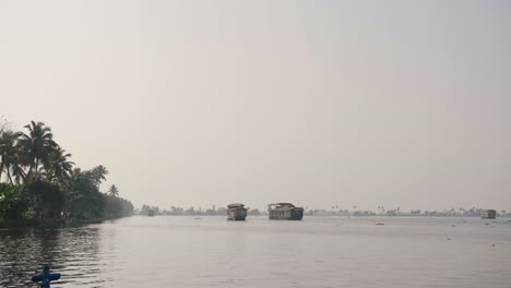 Ein-Offener-Wasserabschnitt-In-Den-Backwaters-Von-Kerala-Zeigt-Mehrere-Hausboote-In-Der-Späten-Nachmittagssonne-Und-Links-Ein-Von-Palmen-Gesäumtes-Ufer
